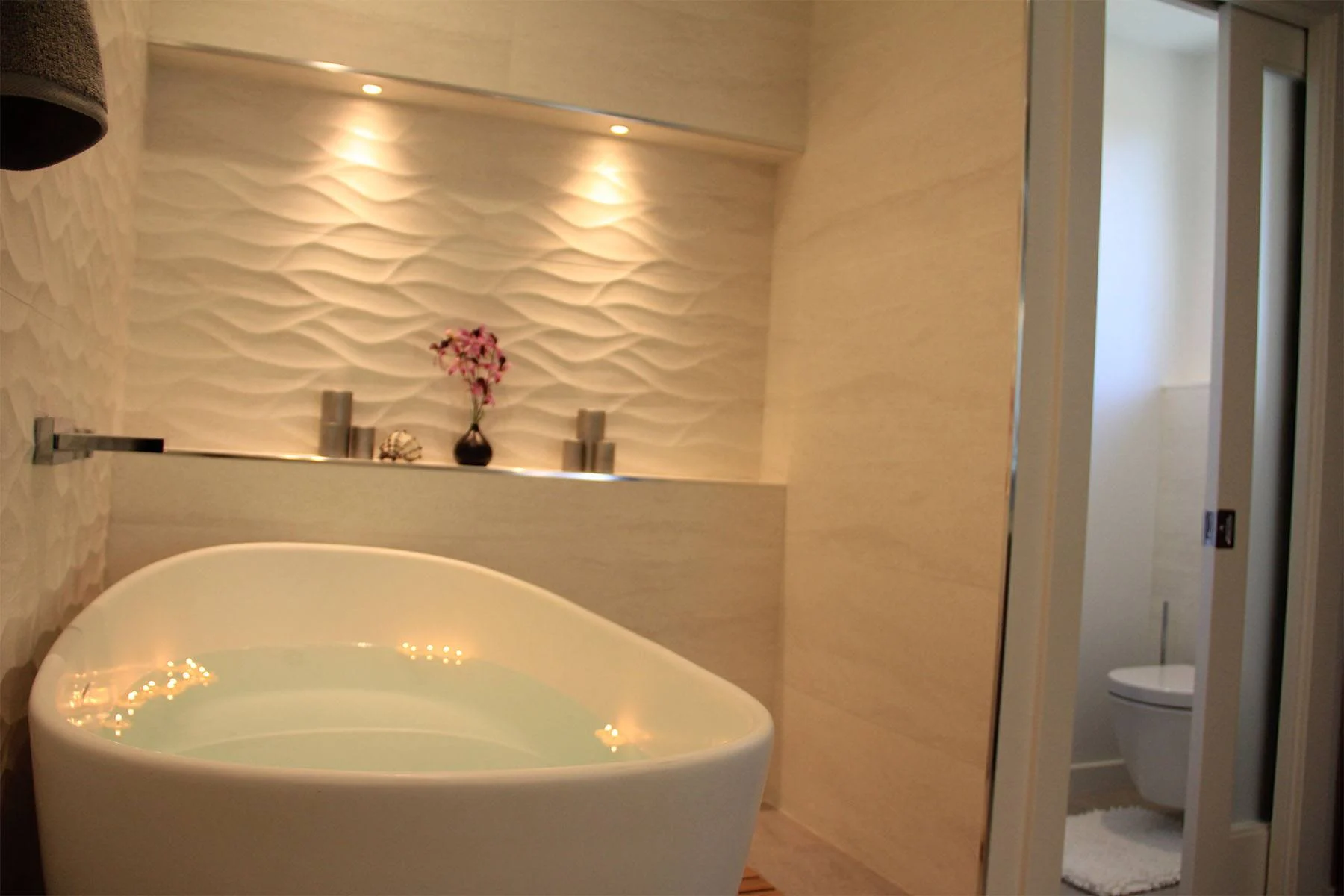 Freestanding soaking tub with lit niche against a wave-textured tile wall