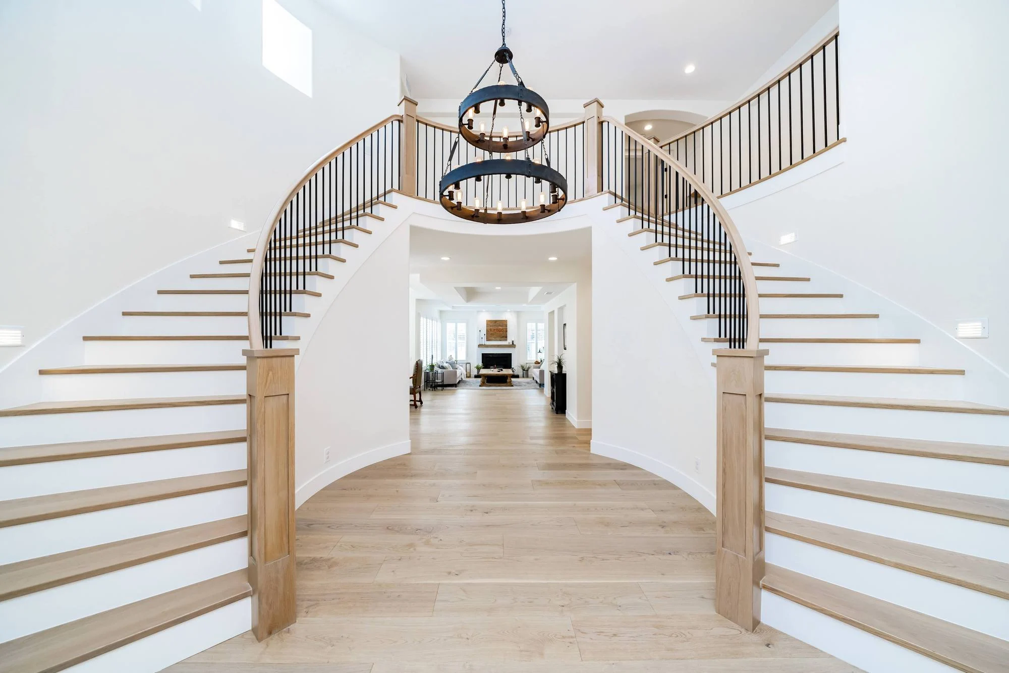 Double curved staircase with iron balusters and round chandelier over a white oak foyer