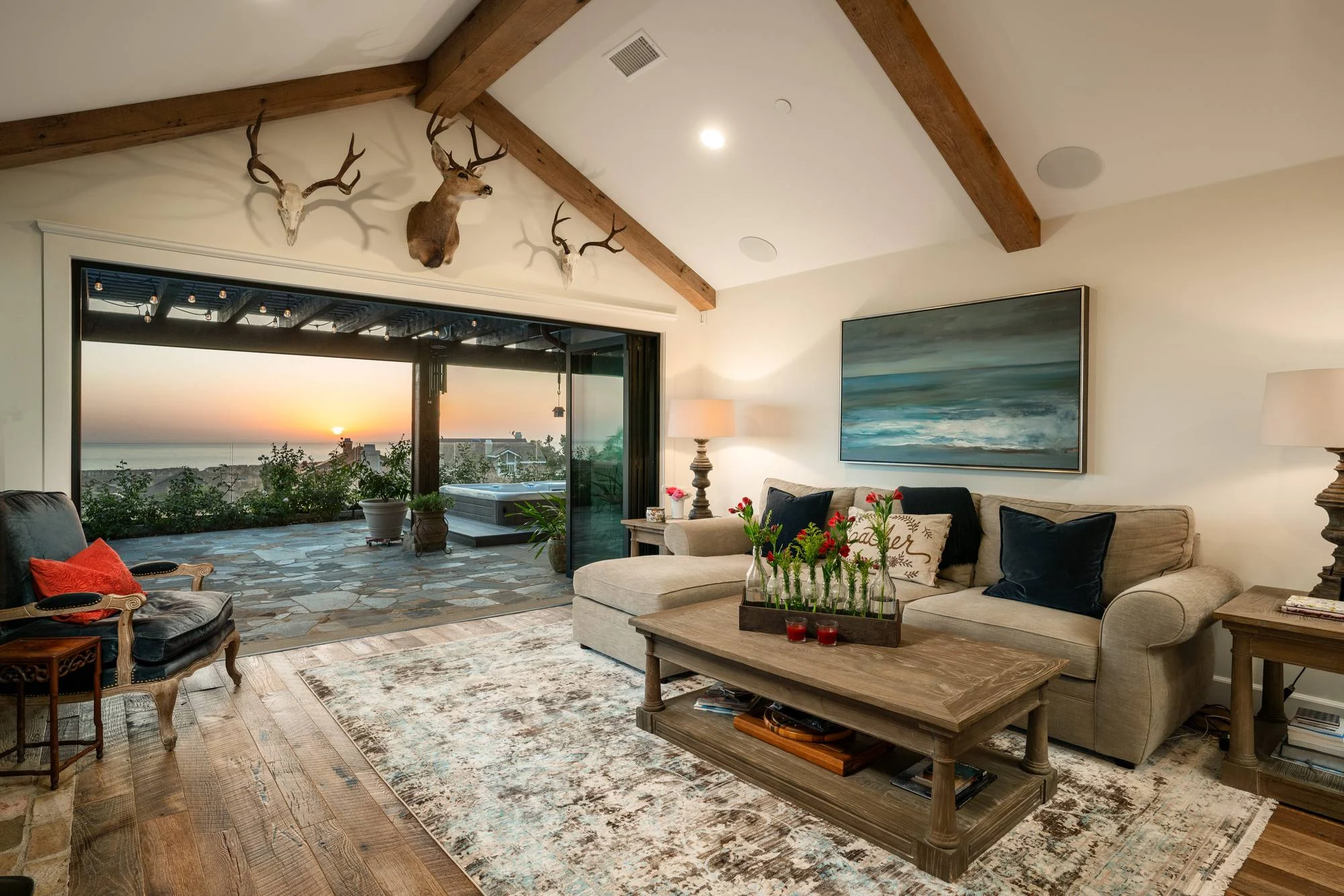 Vaulted living room with exposed beams and wood floors opening through wide glass doors to an ocean sunset