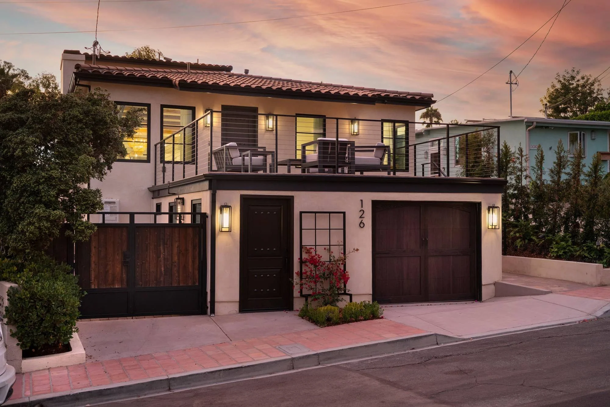 Two-story Spanish-style beach house at sunset with wood garage doors, roof deck and lit sconces