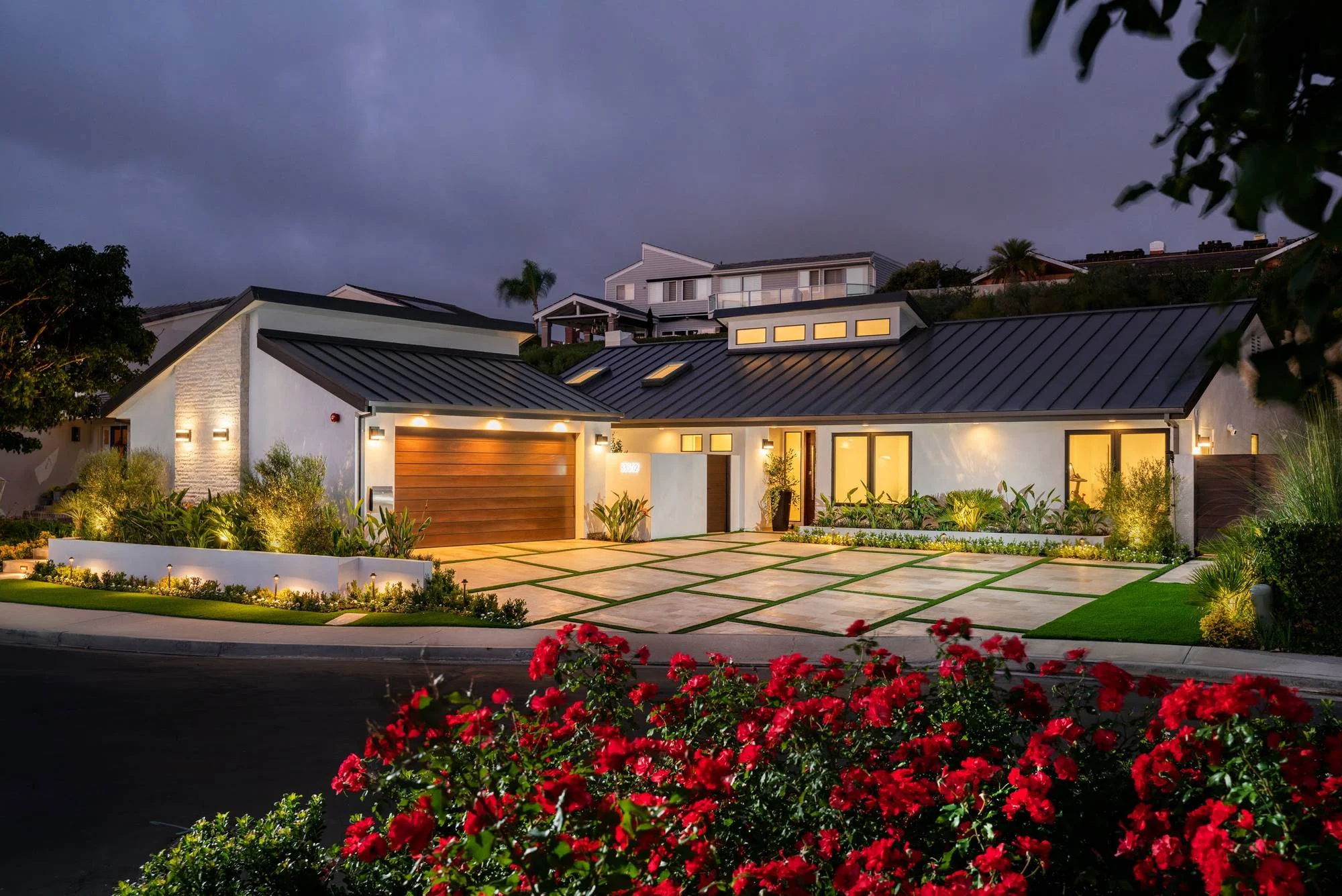 Contemporary hilltop home at dusk with standing-seam roof, wood garage door and paver driveway